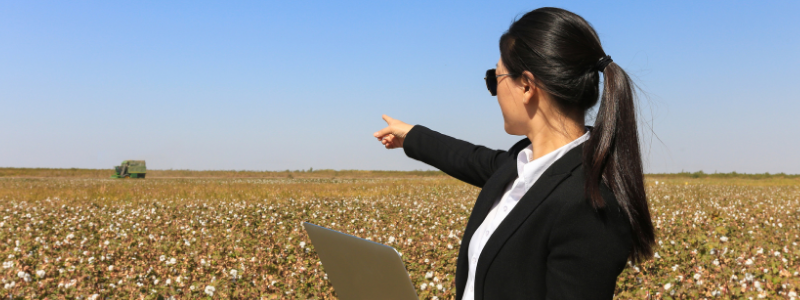 Woman Holding Laptop and Pointing to a Field of Crops